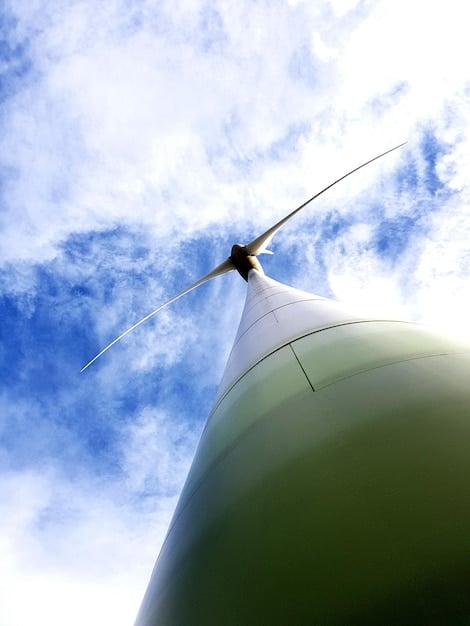 A close-up shot of a modern wind turbine with a vibrant green field in the background and a clear blue sky overhead. The image celebrates the symbiosis between advanced renewable energy technology and the natural environment.