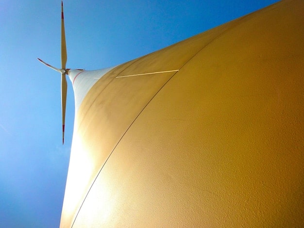 A detailed close-up of a wind turbine in a rural area. The blades are turning slowly, and the sun is setting in the background, creating a warm glow.