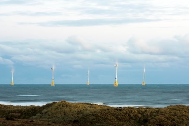 An aerial view of an offshore wind farm during the day. The turbines are evenly spaced and aligned, creating a grid pattern in the ocean. The sea is a deep blue, and the horizon is visible in the distance. A service vessel is approaching one of the turbines, indicating ongoing maintenance and operation.
