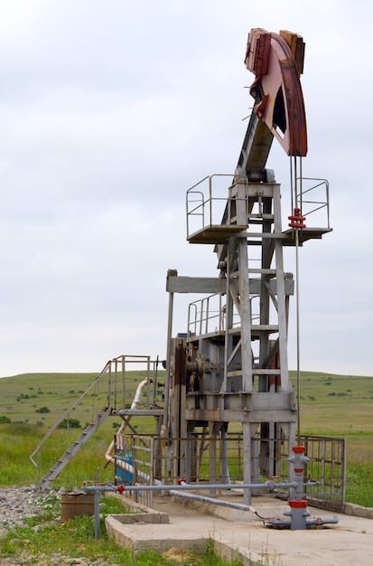 A close-up view of a geothermal drilling rig in operation. The rig is tall and complex, with various pipes, machinery, and a rotating drill bit extending into the ground. The surrounding area is rocky and arid, and the sky is clear. The image highlights the technical complexity and scale of geothermal drilling operations.